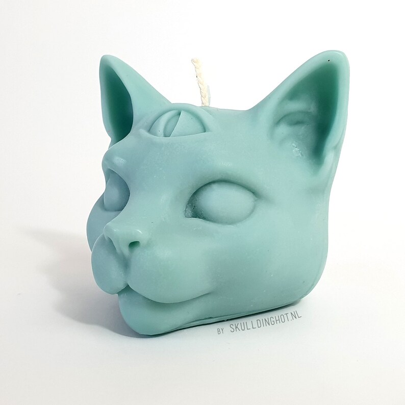 Puede incluir: Una vela de cabeza de gato azul turquesa con un tercer ojo en la frente. La vela tiene una mecha blanca y est&aacute; hecha por SKULLDINGHOT.NL.