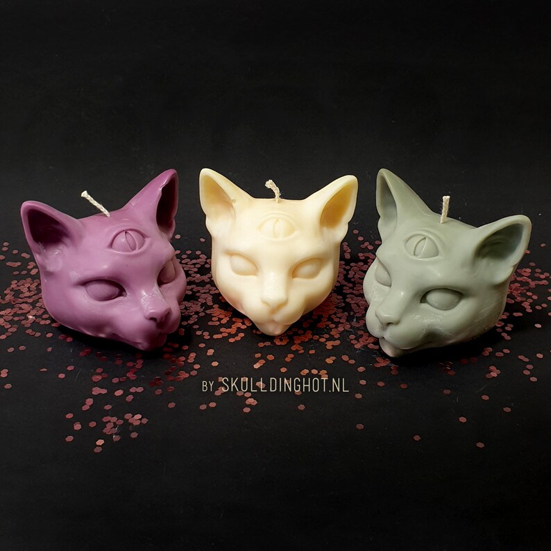 Puede incluir: Tres velas con forma de cabeza de gato, cada una con un tercer ojo, en morado, blanco y verde. Las velas est&aacute;n sobre un lecho de purpurina roja.