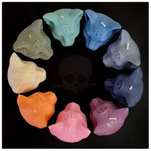 Puede incluir: Un conjunto de diez velas de cabeza de gato de colores, cada una con un solo ojo y un color diferente. Las velas est&aacute;n dispuestas en c&iacute;rculo sobre un fondo negro.