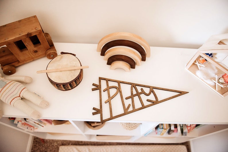 Pu&ograve; includere: Uno scaffale in legno per la sala giochi con una variet&agrave; di giocattoli, tra cui un camion giocattolo in legno, un piccolo tamburo, un arcobaleno in legno e un cartello in legno che dice "PLAY".