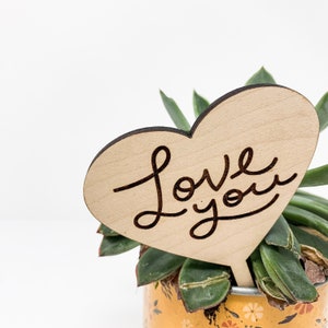 May include: A wooden heart-shaped sign with the words "Love you" written in cursive. The sign is placed in a pot with a succulent plant.