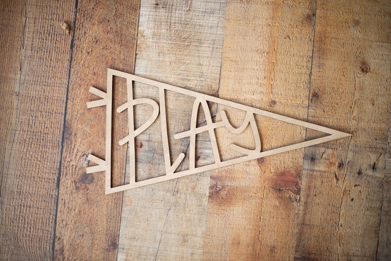 Pu&ograve; includere: Un pennone di legno con la parola "PLAY" tagliata in un carattere fantasioso. Il pennone &egrave; di colore marrone chiaro e si trova su una superficie di legno.