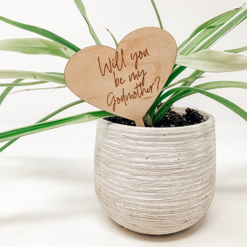 Puede incluir: Un cartel de madera en forma de coraz&oacute;n con el texto "&iquest;Quieres ser mi madrina?" El cartel est&aacute; clavado en la tierra de una planta en maceta.