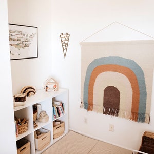 Pu&ograve; includere: Una libreria bianca con vari oggetti, tra cui libri, cesti e giocattoli di legno. Un arazzo da parete intrecciato con colori arcobaleno con frange &egrave; appeso al muro dietro la libreria.