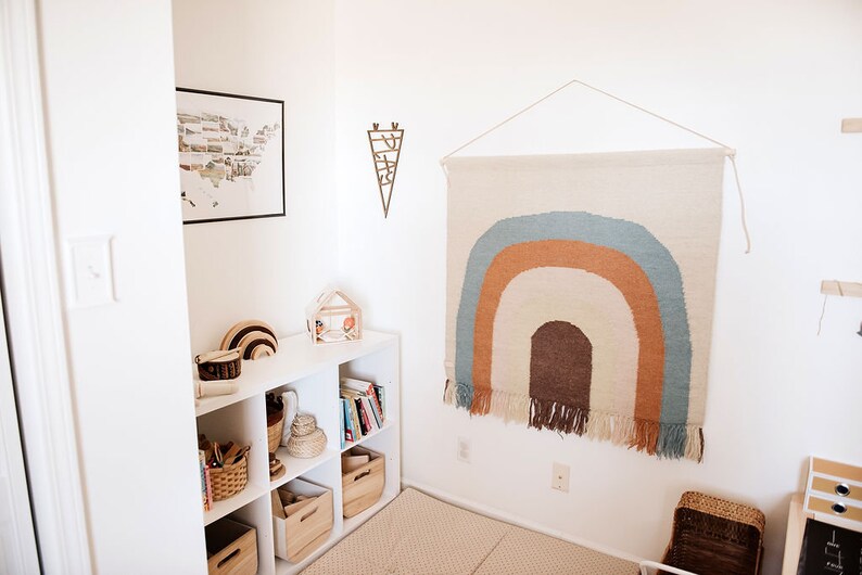 Pu&ograve; includere: Una libreria bianca con vari oggetti, tra cui libri, cesti e giocattoli di legno. Un arazzo da parete intrecciato con colori arcobaleno con frange &egrave; appeso al muro dietro la libreria.