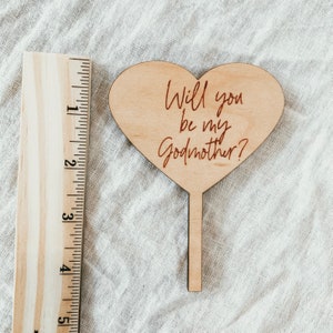 Puede incluir: Un letrero de madera en forma de coraz&oacute;n con el texto "&iquest;Quieres ser mi madrina?" en letras cursivas rojas. El letrero est&aacute; unido a un palo de madera.