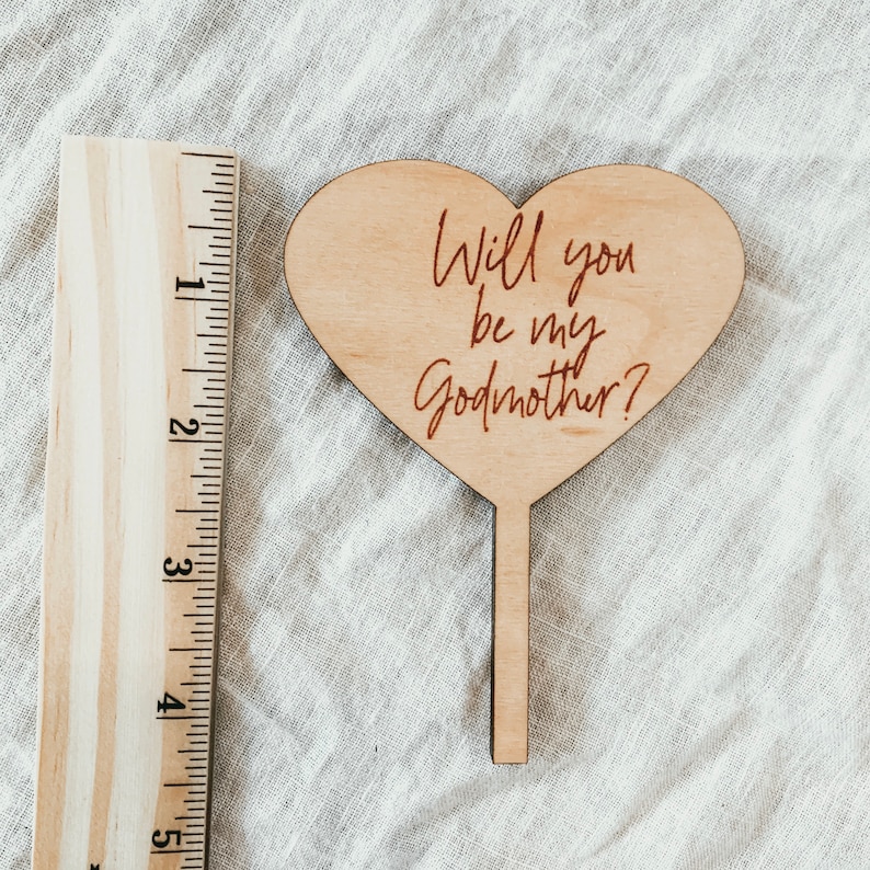 Puede incluir: Un letrero de madera en forma de coraz&oacute;n con el texto "&iquest;Quieres ser mi madrina?" en letras cursivas rojas. El letrero est&aacute; unido a un palo de madera.