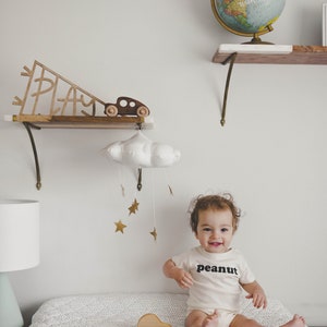 Pu&ograve; includere: Un bambino indossa un body bianco con la scritta "peanut" stampata su di esso, &egrave; seduto su un fasciatoio con un motivo a pois bianco e nero. Un'auto giocattolo di legno &egrave; sul fasciatoio. Un mobile a forma di nuvola bianca con stelle dorate &egrave; appeso sopra il bambino.