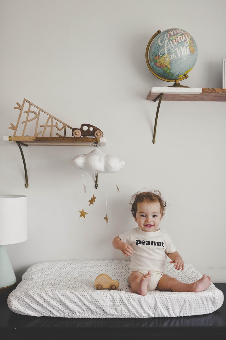 Pu&ograve; includere: Un bambino indossa un body bianco con la scritta "peanut" stampata su di esso, &egrave; seduto su un fasciatoio con un motivo a pois bianco e nero. Un'auto giocattolo di legno &egrave; sul fasciatoio. Un mobile a forma di nuvola bianca con stelle dorate &egrave; appeso sopra il bambino.