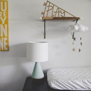 Pu&ograve; includere: Una lampada bianca con una base blu chiaro si trova su una cassettiera nera con un fasciatoio bianco in cima. Uno scaffale in legno con la parola "PLAY" ritagliata &egrave; appeso al muro sopra la cassettiera. Un mobile a forma di nuvola bianca con stelle dorate &egrave; appeso allo scaffale.