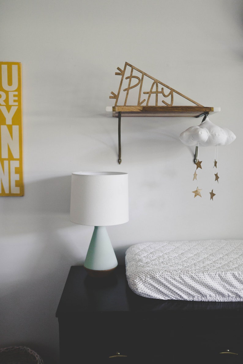 Pu&ograve; includere: Una lampada bianca con una base blu chiaro si trova su una cassettiera nera con un fasciatoio bianco in cima. Uno scaffale in legno con la parola "PLAY" ritagliata &egrave; appeso al muro sopra la cassettiera. Un mobile a forma di nuvola bianca con stelle dorate &egrave; appeso allo scaffale.