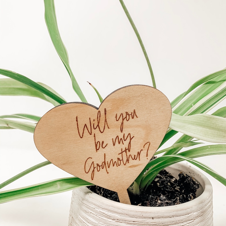 Puede incluir: Un letrero de madera en forma de coraz&oacute;n con el texto "&iquest;Quieres ser mi madrina?" escrito en cursiva. El letrero est&aacute; colocado en una planta en maceta.