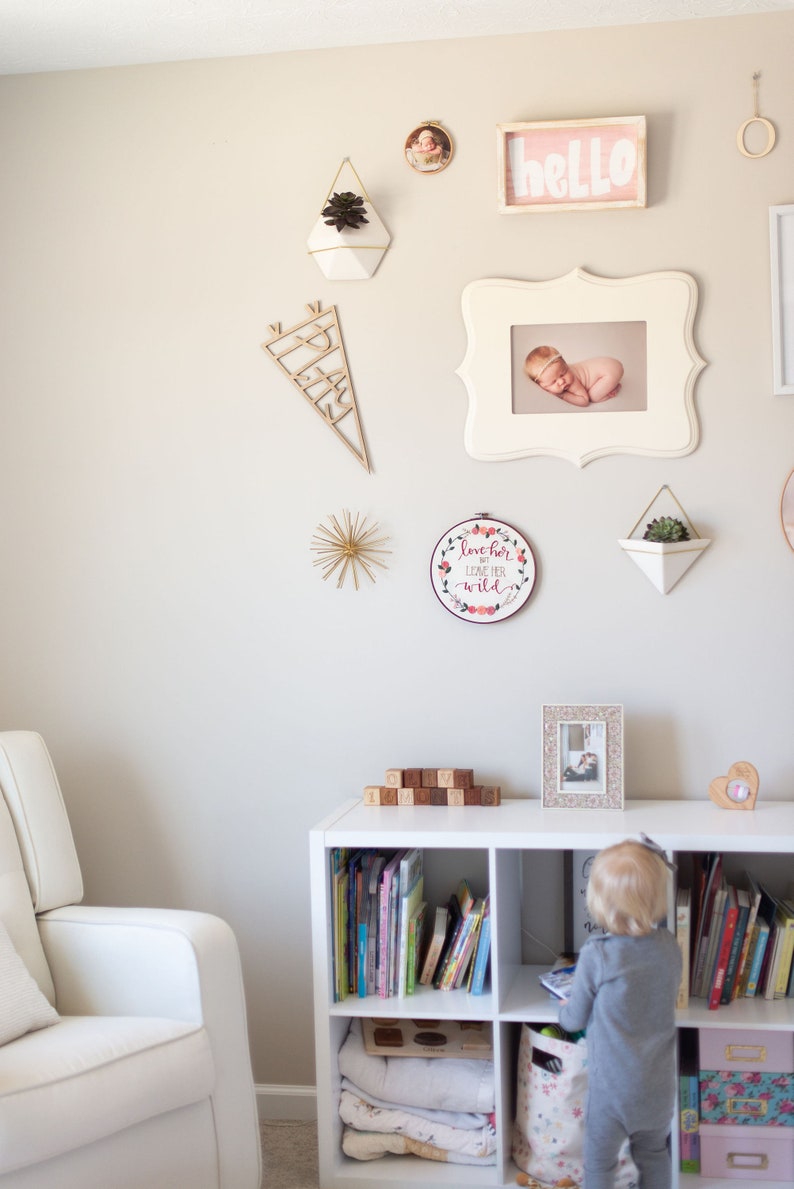 Pu&ograve; includere: Una libreria bianca con libri, coperte e un cesto. Un bambino &egrave; in piedi davanti alla libreria, guardando i libri. La parete dietro la libreria &egrave; decorata con una variet&agrave; di immagini incorniciate, tra cui una foto di un bambino, una foto di una pianta e un cartello che dice "hello".