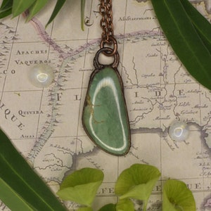 Puede incluir: Un collar de cobre con un colgante de piedra verde y blanca. El colgante está engastado en un marco de cobre y cuelga de una cadena de cobre. El collar está fotografiado sobre un mapa antiguo.