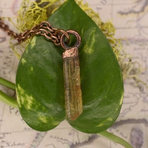 Puede incluir: Un collar de cadena de cobre con un colgante que presenta un cristal largo, delgado, transparente y naranja. El cristal está engastado en un bisel de cobre y cuelga de una anilla de cobre.