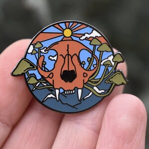 Puede incluir: Un pin de esmalte circular con una calavera de animal roja y naranja rodeada de setas y un cielo azul con un sol amarillo. El pin tiene un contorno blanco y está sobre un fondo azul.