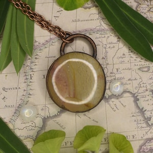 Puede incluir: Un collar de cadena de cobre con un colgante redondo con una piedra de ágata marrón claro con un patrón de círculo blanco. El colgante está sobre un fondo de mapa vintage.