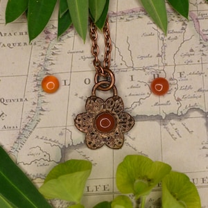 Puede incluir: Un collar con un colgante de flor de cobre con una piedra de cornalina roja en el centro. El collar está sobre un mapa vintage con hojas verdes.