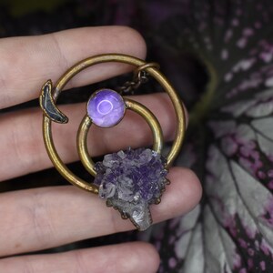 Puede incluir: Un colgante de latón con un cristal de amatista púrpura, un amuleto de luna creciente y una piedra púrpura más pequeña en el centro.