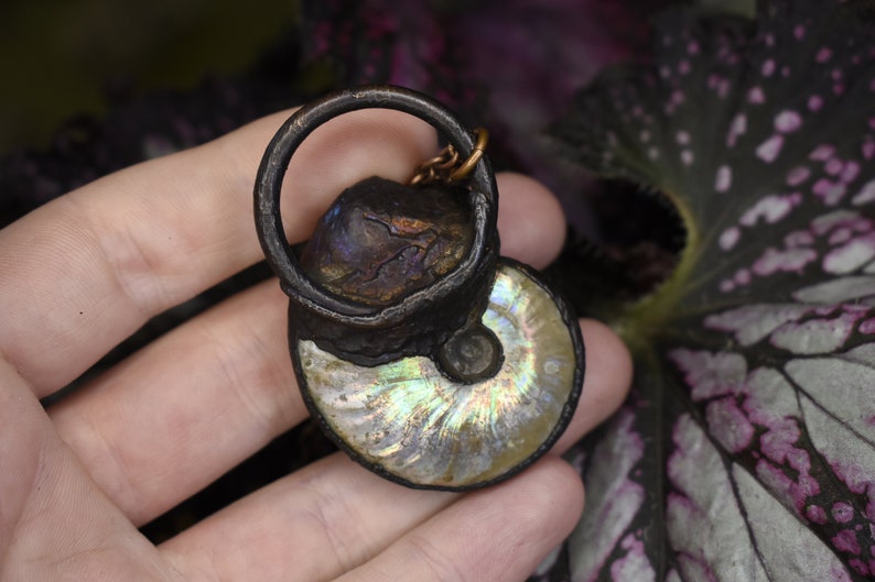 Puede incluir: Un colgante de f&oacute;sil de amonita plateado e iridiscente con un anillo y una cadena de bronce. El colgante se sostiene en una mano sobre un fondo oscuro.