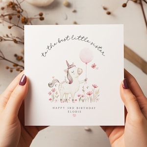 K&ouml;nnte beinhalten: Ein wei&szlig;es Einhorn mit einem rosafarbenen Horn und einem rosafarbenen Ballon im Huf steht auf einer Wiese mit rosa Blumen. Die Karte lautet "an die beste kleine Schwester" und "Happy 3rd Birthday Elodie".