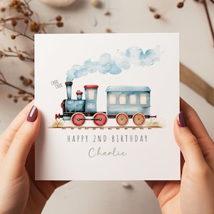 May include: A white birthday card featuring a blue watercolour train with red wheels and a red roof. The card reads "Happy 2nd Birthday Charlie" in black text, with "Choo Choo" above the train.