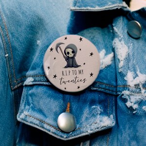 May include: A round button with a cartoon grim reaper holding a scythe. The button has a white background with black stars and the text "R.I.P. TO MY TWENTIES".