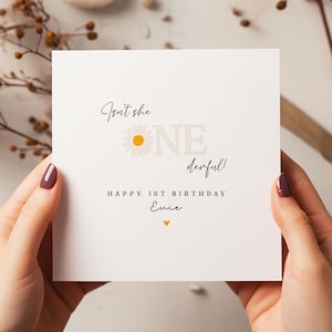 Puede incluir: Tarjeta de cumplea&ntilde;os blanca con el texto "Isn't she ONE derful!" y "HAPPY 1ST BIRTHDAY Evie" en escritura negra. Un gr&aacute;fico de margarita est&aacute; al lado de la palabra "ONE". Un peque&ntilde;o coraz&oacute;n amarillo est&aacute; en la parte inferior.