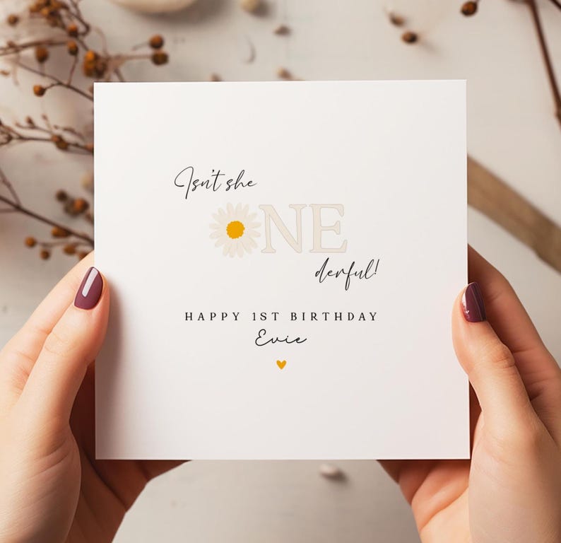 Puede incluir: Tarjeta de cumplea&ntilde;os blanca con el texto "Isn't she ONE derful!" y "HAPPY 1ST BIRTHDAY Evie" en escritura negra. Un gr&aacute;fico de margarita est&aacute; al lado de la palabra "ONE". Un peque&ntilde;o coraz&oacute;n amarillo est&aacute; en la parte inferior.