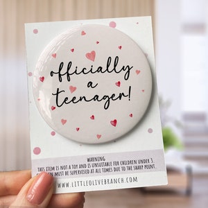 May include: A white button with pink hearts and the text "officially a teenager!" on it. The button is on a white card with pink polka dots and a warning label.