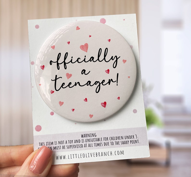 May include: A white button with pink hearts and the text "officially a teenager!" on it. The button is on a white card with pink polka dots and a warning label.