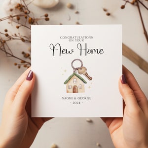 K&ouml;nnte beinhalten: Quadratische Karte mit dem Text "CONGRATULATIONS ON YOUR New Home". Sie zeigt eine Illustration eines Hauses und eines Schl&uuml;ssels mit den Namen "NAOMI & GEORGE" und dem Jahr "2024".