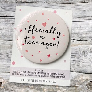 May include: A white round button with a pink background and red hearts. The button says "officially a teenager!" in black text.