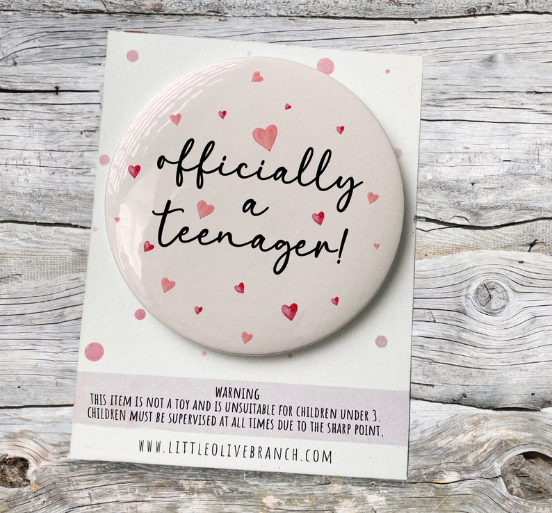 May include: A white round button with a pink background and red hearts. The button says "officially a teenager!" in black text.
