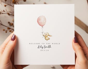May include: A white card with a pink balloon with the number 2024 on it. A small bunny is holding onto the balloon string. The text "Welcome to the world Lily Smith 28.11.24" is printed on the card.