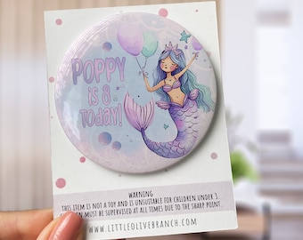 May include: A purple and blue mermaid with a crown and a purple tail holds balloons that say "Poppy is 8 Today!" on a round button.