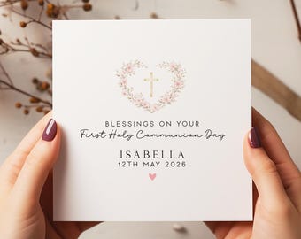 May include: A white card with the text "Blessings on Your First Holy Communion Day" and the name "Isabella" with the date "12th May 2026". The card features a gold cross inside a floral heart design and a small pink heart.