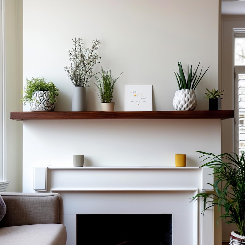 Puede incluir: Una chimenea blanca con una repisa de madera oscura exhibe varias plantas en macetas y objetos decorativos. Un jarr&oacute;n gris, una maceta blanca en forma de pi&ntilde;a y una peque&ntilde;a maceta negra se encuentran entre los objetos. Tambi&eacute;n hay una tarjeta de felicitaci&oacute;n con texto.