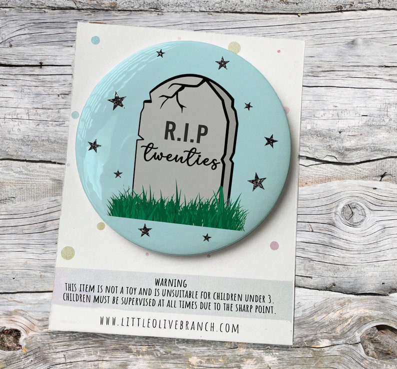 May include: A light blue round button with a black and white illustration of a tombstone with the text "R.I.P twenties" on it. The tombstone is surrounded by green grass and black stars.
