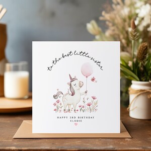 K&ouml;nnte beinhalten: Ein wei&szlig;es Einhorn mit einem rosafarbenen Horn und einem rosafarbenen Ballon in seinen Hufen steht auf einer Wiese mit rosa und gr&uuml;nen Blumen. Der Text "to the best little sister" ist in einer kursivschriftlichen Schriftart &uuml;ber dem Einhorn geschrieben. Der Text "Happy 3rd Birthday Elodie" ist unter dem Einhorn geschrieben.