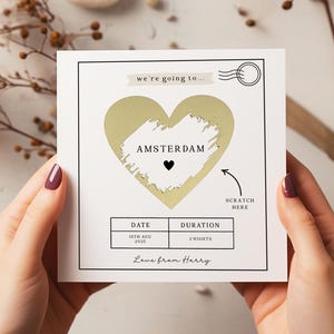 May include: A white card with a gold heart-shaped scratch-off area revealing "AMSTERDAM" and a black heart. The card includes the text "we're going to...", "DATE", "DURATION", and "Love from Harry". A scratch-off arrow is visible.