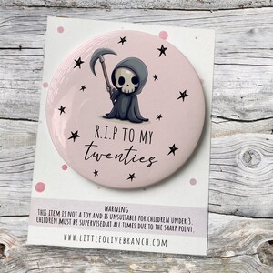 May include: A pink round mirror with a cartoon image of the Grim Reaper holding a scythe. The mirror has a white border and the text "R.I.P TO MY twenties" in black. The background of the mirror is pink with white stars.