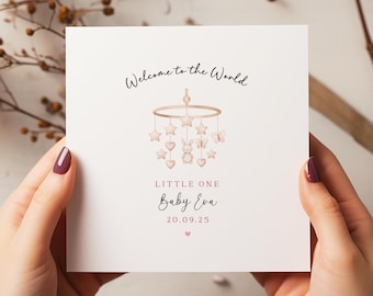 May include: A square, white card with the text "Welcome to the World" and "Little One Baby Eva 20.09.25". The card features a baby mobile design with stars, hearts, and a bunny. The card is held by two hands.