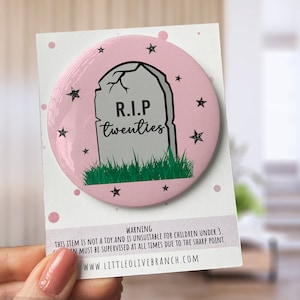 May include: A pink button with a white tombstone that says "R.I.P. twenties" and green grass at the base. The button is surrounded by white stars on a pink background.