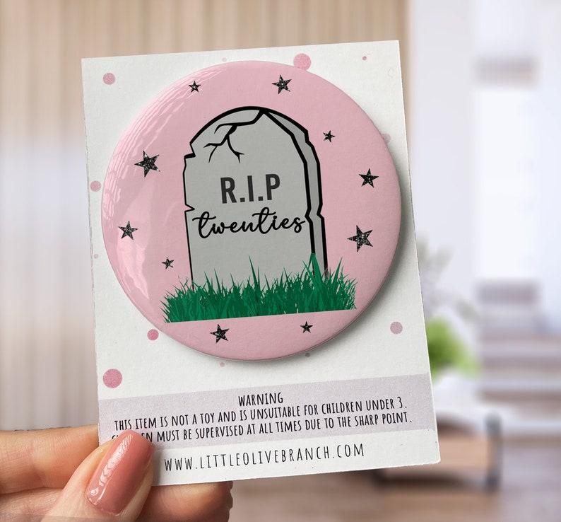 May include: A pink button with a white tombstone that says "R.I.P. twenties" and green grass at the base. The button is surrounded by white stars on a pink background.