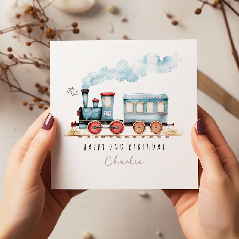 May include: A white birthday card featuring a blue watercolour train with red wheels and a red roof. The card reads "Happy 2nd Birthday Charlie" in black text, with "Choo Choo" above the train.