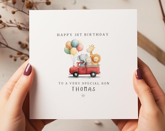 May include: A white greeting card with the text "HAPPY 1ST BIRTHDAY" and "TO A VERY SPECIAL SON THOMAS". The card features a red truck carrying animal illustrations and balloons.