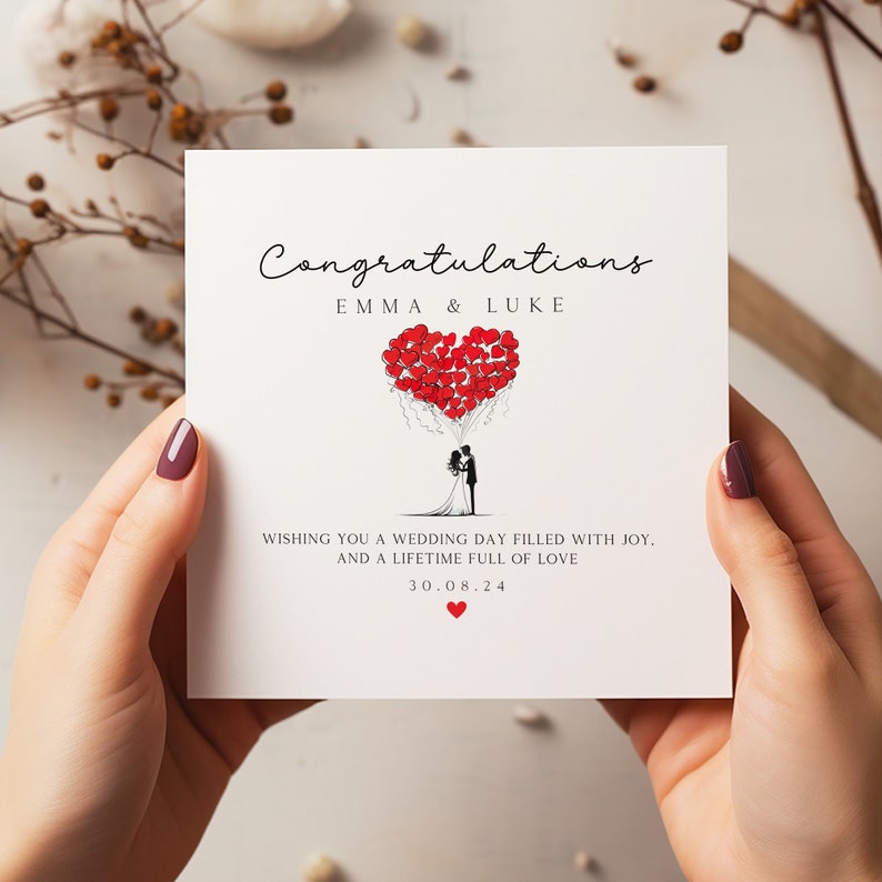 May include: A white card with black text that reads "Congratulations Emma & Luke" and "Wishing you a wedding day filled with joy, and a lifetime full of love 30.08.24". A black and white illustration of a bride and groom standing under a heart-shaped balloon made of red hearts.
