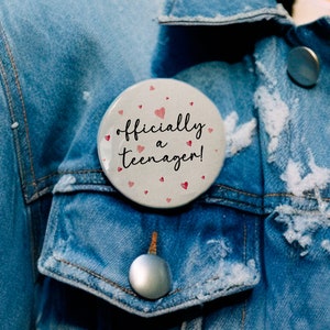 May include: A white button with pink hearts and the text "officially a teenager!" on a blue denim jacket.