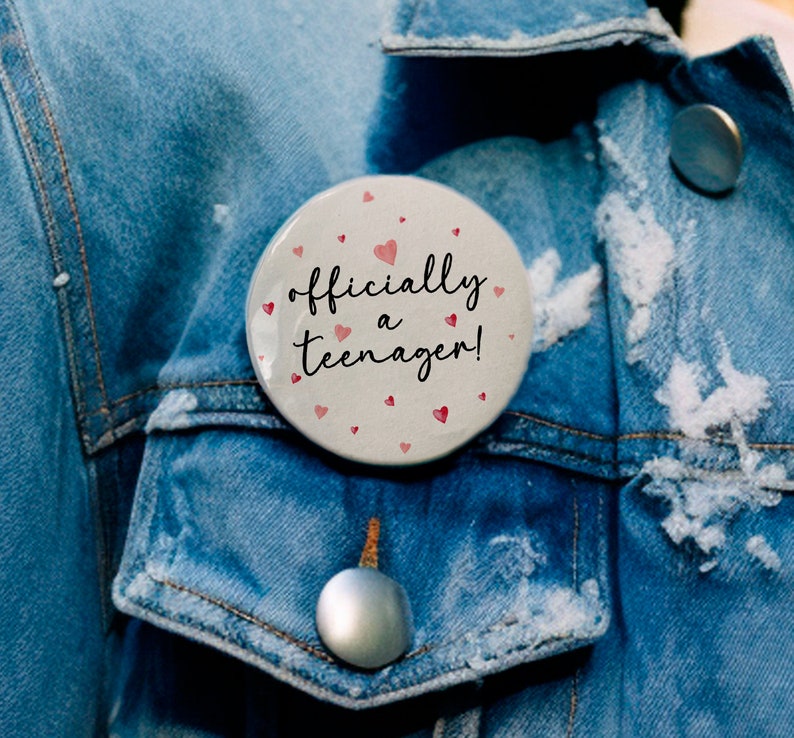 May include: A white button with pink hearts and the text "officially a teenager!" on a blue denim jacket.